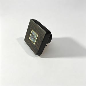 May include: A square cabinet knob featuring a dark brown wood grain finish with a central square inlay of iridescent abalone shell framed by a thin gold-coloured metal border. The knob has a smooth, rounded edge profile and a cylindrical base.