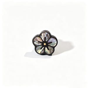 May include: A flower-shaped button featuring a dark wood frame with five petals inlaid with iridescent abalone shell. The centre of the floral design is accented with a small, circular gold-toned metal stud, creating a contrast between the dark wood and the shimmering shell material.