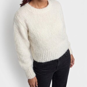 Könnte beinhalten: Ein cremefarbener, langärmeliger, flauschiger Strickpullover mit Rundhalsausschnitt sowie gerippten Bündchen an Ärmeln und Saum, kombiniert mit einer schwarzen Jeans. Der Pullover hat eine weiche, strukturierte Optik und eine kurze Passform.