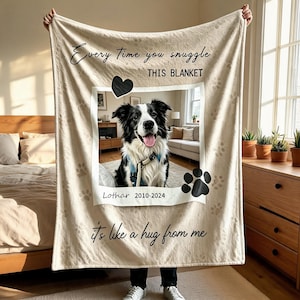 Op de afbeelding: Een beige deken met een centraal fotokader met een zwart-witte border collie, de tekst "Every Time you snuggle THIS BLANKET it's like a hug from me", de naam "Lothar 2010-2024" en zwarte pootafdrukken als decoratieve accenten.