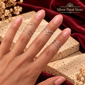 May include: A set of silver-toned rings worn on fingers, featuring three thin bands on the index finger and two thin bands on the middle finger. Some rings have a V-shaped chevron design, while others are simple circular bands. Silver Patal Store.