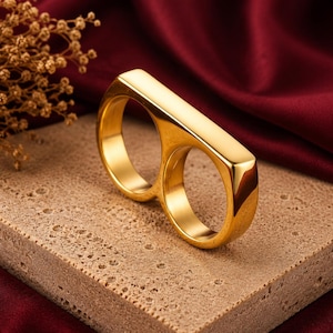 May include: A polished gold-tone double finger ring featuring a smooth, elongated rectangular bar connecting two circular bands. The ring has a reflective, metallic finish and a solid, continuous construction designed to be worn across two adjacent fingers.