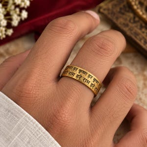 May include: A gold-toned band ring featuring black engraved Sanskrit text that reads Hare Krishna Hare Krishna Hare Rama Hare Rama. The ring has a matte, textured finish and a flat profile, encircling the finger with clear, legible lettering.