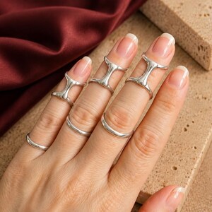 May include: A set of silver-toned rings worn on the fingers. Three rings feature a distinctive H-shaped design that spans the middle knuckle, while three plain, smooth, thin bands are positioned at the base of the fingers on the hand.