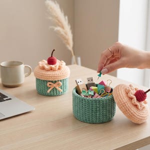 May include: Two round, crocheted containers shaped like cupcakes with mint green bases and peach-colored tops. Each lid features a small, dark red crocheted cherry. One container is open, revealing various office supplies like USB drives and clips.
