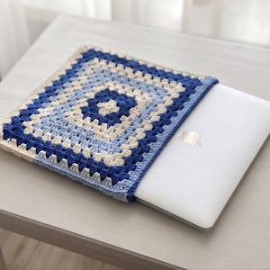 Puede incluir: Una funda de ganchillo cuadrada para portátil con un patrón de granny square en tonos azul oscuro, azul claro y crema. La funda está confeccionada con un punto de hilo texturizado y contiene un portátil plateado con el logo de apple.