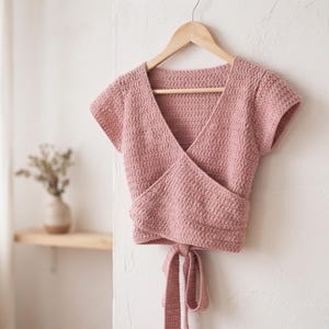 May include: A dusty rose crochet wrap top with short sleeves, a deep V-neckline, and a tie-front closure. The garment is crafted with a textured stitch pattern and is displayed on a wooden hanger against a plain, light-colored wall surface.