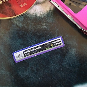 May include: A rectangular sticker featuring a digital music player interface with the text "Freak On A Leash" and "Korn" displayed. The sticker has a purple border with a subtle star pattern, a pause icon, and green equalizer bars.
