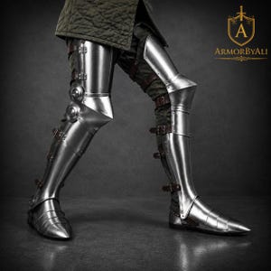 May include: Polished steel leg armor featuring articulated greaves, poleyns, and cuisses secured with brown leather straps and metal buckles. The metallic plating covers the lower limbs, extending from the upper thigh down to the pointed sabatons.