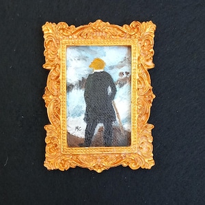 May include: A miniature rectangular painting in an ornate gold-coloured frame. The artwork depicts a figure with blonde hair seen from behind, wearing a dark coat and standing in a snowy landscape. The initials MC are visible in the bottom left.