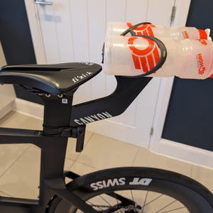 May include: A black Canyon bicycle seat post and saddle with a fi'zi:k brand label. Attached to the rear of the saddle is a black dual bottle cage mount holding two clear plastic water bottles with red caps and High5 branding in red text.