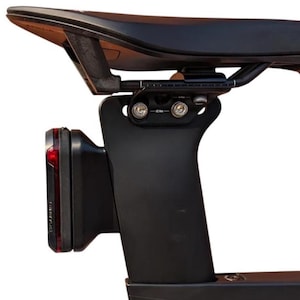 May include: A black bicycle seat post and saddle assembly featuring a mounted red Garmin rear light. The hardware includes a metal bracket secured with two bolts marked 10 Nm, attached to the underside of the saddle rails with a visible measurement scale.