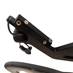Pode incluir: Um componente de bicicleta em metal preto com um braço retangular, dois insertos roscados na cor latão e dois parafusos sextavados prateados. Um cordão elástico preto com um pequeno fecho de plástico preto está preso na extremidade.