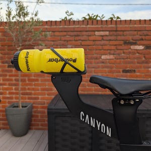May include: A bright yellow Powerbar water bottle secured in a black bottle cage mounted behind a black bicycle saddle. The bicycle frame is black with the white text CANYON printed on the side. The setup is positioned for cycling hydration.