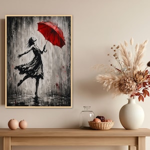 May include: A framed wall art print featuring a black and white ink-style illustration of a person in a dress dancing in the rain while holding a vibrant red umbrella. The artwork uses high-contrast splatters and drips to depict a rainy scene.