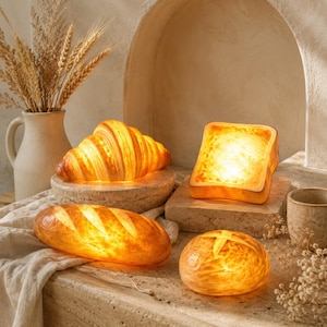 May include: A collection of four bread-shaped lamps emitting a warm glow, including a croissant, a slice of toast, a baguette, and a round loaf. The lamps are arranged on stone surfaces, with a ceramic pitcher and dried wheat stalks nearby.