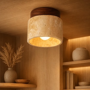 May include: A flush mount ceiling light fixture featuring a cylindrical shade made of porous beige travertine stone, topped with a dark brown wooden circular base, emitting a warm glow from an internal light bulb directed downwards.