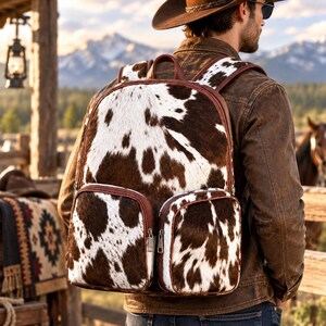 Puede incluir: Una mochila con estampado de piel de vaca en marrón y blanco, que cuenta con un compartimento principal grande y dos bolsillos delanteros más pequeños, asegurados con ribetes de cuero sintético marrón y cremalleras metálicas.