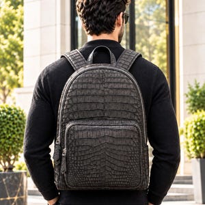 May include: A dark charcoal grey backpack featuring a textured crocodile skin pattern. The bag includes a large main compartment with a silver zip, a smaller front pocket with dual zip pulls, a top handle, and two adjustable shoulder straps.