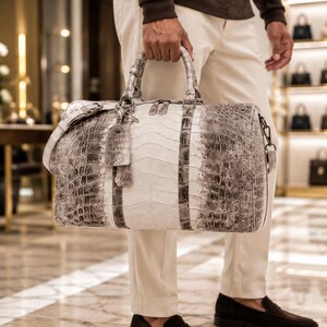 May include: A large duffel bag featuring a textured, scale-like pattern in shades of white, grey, and charcoal. The bag includes two top handles, a detachable shoulder strap, a hanging luggage tag, and a dual-zipper closure across the top.