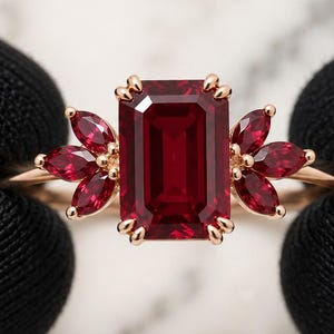 Könnte beinhalten: Ein Goldring mit einem großen, smaragdgeschliffenen, tiefroten Edelstein in der Mitte, flankiert von drei kleineren, marquise-geschliffenen roten Edelsteinen auf jeder Seite. Die Fassung besteht aus poliertem Gold und betont die leuchtenden Steine.