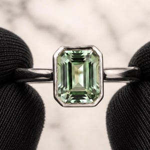 Peut inclure: Une bague ornée d'une pierre précieuse vert clair de taille émeraude rectangulaire, sertie dans un chaton en métal argenté poli. La pierre présente une surface facettée claire avec une profondeur interne visible, montée sur un anneau fin.