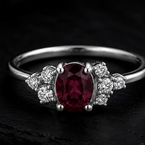 Può includere: Un anello color argento con una pietra preziosa centrale rosso scuro dal taglio ovale. La pietra è affiancata su entrambi i lati da un gruppo di tre piccole pietre bianche rotonde e chiare su una fascia metallica, creando un gioiello dettagliato.