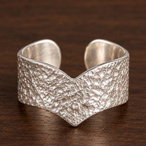 Può includere: Un anello aperto color argento caratterizzato da una punta a forma di V al centro. La fascia ha un profilo largo e piatto con una finitura superficiale testurizzata e martellata che crea un aspetto ruvido e irregolare su tutta la superficie.