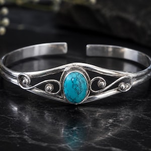 May include: A silver cuff bracelet featuring a central oval turquoise stone with dark veining. The metal band displays a polished finish and decorative swirling wirework details on both sides of the stone, creating an open, symmetrical design.