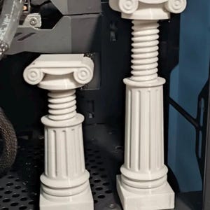 May include: Two light grey, 3D-printed decorative columns featuring a square base, a fluted lower section, a ribbed middle section, and a volute capital top. The columns are positioned upright, with one being taller than the other, displaying a smooth, matte finish and architectural design elements.