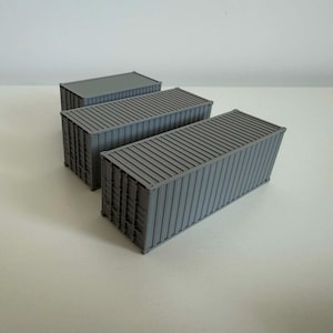 May include: Three miniature grey shipping containers with corrugated walls and rectangular roofs. Each model features vertical ribbing along the sides and ends, mimicking the appearance of standard industrial freight containers in a small scale.