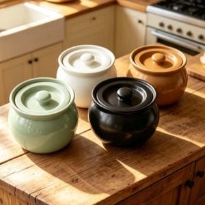 Może przedstawiać: Cztery okrągłe ceramiczne słoiki do przechowywania z pasującymi pokrywkami, ustawione w grupie. Zestaw zawiera słoik jasnozielony, biały, czarny i jasnobrązowy. Każdy pojemnik ma gładkie wykończenie i mały uchwyt w kształcie gałki na pokrywce.