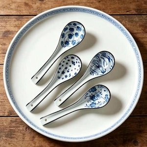 May include: Four white ceramic spoons with blue patterns arranged on a large white plate with a blue rim. Each spoon features a unique blue design, including floral motifs, small dots, and wave-like patterns, with a small hole at the handle end.