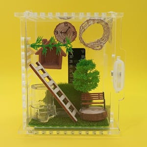 May include: A clear acrylic enclosure for small animals featuring a miniature landscape with a wooden ladder, a brown bench, a green tree, a small house, a round clock, a circular rope ring, and a black thermometer displaying temperature in Fahrenheit.
