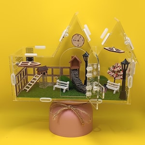May include: A clear acrylic house-shaped enclosure featuring a miniature park scene with green turf, two white benches, black lampposts, a stone staircase, a small wooden house, a ladder, and a flowering tree. A clock is printed on the back wall.