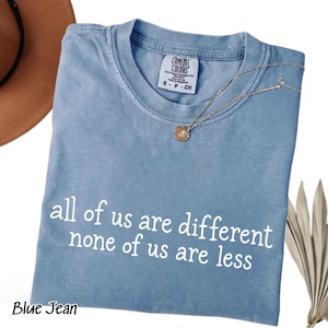May include: A blue jean colored t-shirt with a crew neckline and a white graphic print that reads all of us are different none of us are less. The shirt features a Comfort Colors brand label at the inner neck and a thin gold tone necklace.