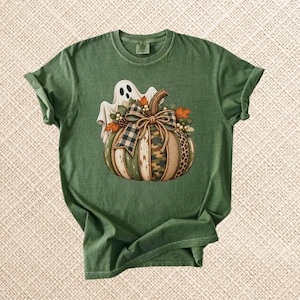 Può includere: Una t-shirt a maniche corte verde oliva con la grafica di un fantasma bianco dietro una zucca. La zucca è decorata con un fiocco a quadri, foglie autunnali e segmenti con motivi mimetici, leopardati e verde tinta unita.