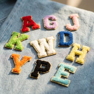 May include: A collection of eleven individual varsity-style letter patches featuring a textured, fuzzy chenille surface with gold-toned metallic trim. The letters include red A, purple G, pink J, green K, white W, blue D, yellow H, orange Y, black P, and light blue E.