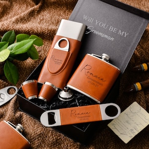 May include: A black gift box containing a tan leather-wrapped flask, a cigar case with a cutter, two small cups, a funnel, and a bottle opener. The items are engraved with the name Romain, Groomsman, and the date 07.09.25. The box lid reads WILL YOU BE MY Groomsman.