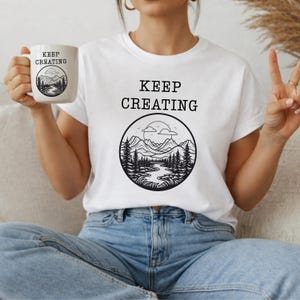 May include: A white t-shirt and a white ceramic mug, both featuring the black text KEEP CREATING above a circular graphic illustration of a mountain range, a forest, a river, and a sun in the sky, all rendered in a black line art style.