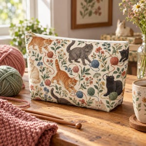 May include: A zippered pouch featuring a cream-colored fabric printed with a pattern of various cats, colorful yarn balls, and floral botanical illustrations. The pouch has a gold-toned metal zipper pull and a structured, rectangular shape.