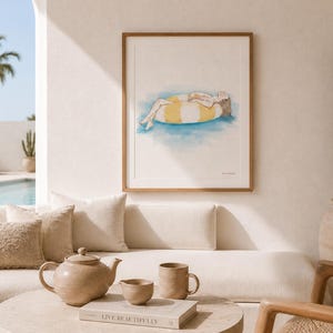 Peut inclure: Une impression d'art encadrée représentant une illustration à l'aquarelle d'une personne allongée sur une bouée rayée jaune et blanche dans une eau bleue. L'œuvre est présentée dans un cadre en bois clair sur un mur neutre au-dessus d'un canapé.