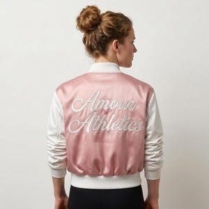 May include: A satin bomber jacket featuring a dusty pink back panel and white sleeves. The back displays the white embroidered text Amour Athletics in a cursive script. The jacket includes a white ribbed collar, cuffs, and a hem at the waist.