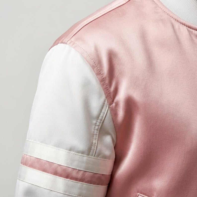 May include: A satin bomber jacket featuring a dusty pink body and contrasting white sleeves. The sleeves are accented with two horizontal white stripes near the cuff area. The garment has a round neckline and visible stitching along the shoulder seams.