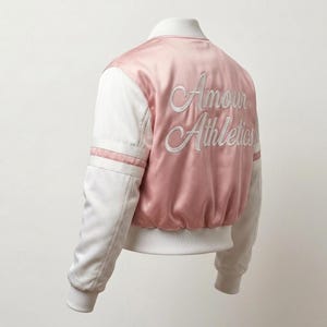 May include: A satin bomber jacket featuring a pale pink back panel with the white embroidered text Amour Athletics in a cursive script. The jacket has white sleeves with two horizontal pink stripes, a white ribbed collar, and a white ribbed hem.