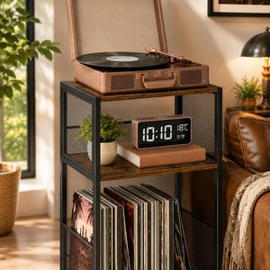 May include: A three-tier storage stand with a black metal frame and dark wood shelves. The top shelf holds a rose gold suitcase-style record player. The middle shelf features a small potted plant, a book, and a digital clock displaying 10:10.