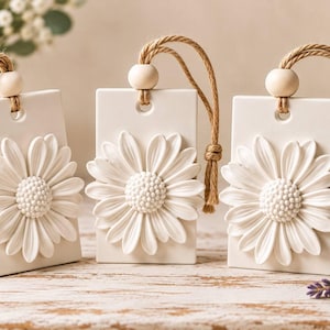 May include: Three rectangular white ceramic plaques, each featuring a raised white daisy flower relief. Each plaque is suspended by a tan jute rope threaded through a hole at the top, accented with a single round wooden bead.