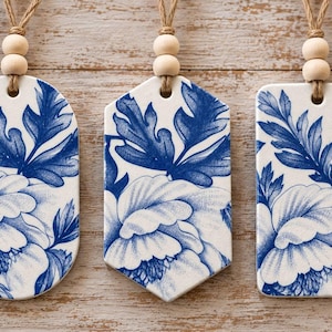 May include: Three ceramic hanging ornaments in oval, hexagonal, and rectangular shapes. Each piece features a white base with a blue floral pattern, two small wooden beads on the top, and a brown twine loop for hanging purposes.