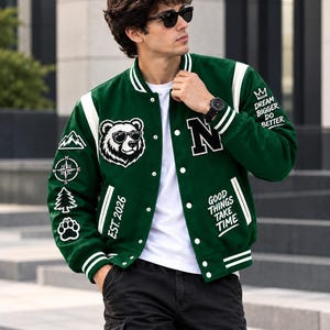 Puede incluir: Una chaqueta universitaria verde oscuro con bordes acanalados a rayas blancas, con un oso con gafas de sol, una brújula, una montaña, un pino y una huella. El texto incluye "N", "EST. 2026", "GOOD THINGS TAKE TIME" y "DREAM BIGGER DO BETTER".