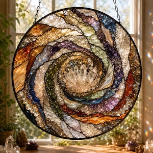 May include: A circular stained glass suncatcher featuring a swirling galaxy design with a central cluster of clear quartz crystals. The piece incorporates textured glass in shades of amber, purple, blue, green, and white, framed in a dark metal border.